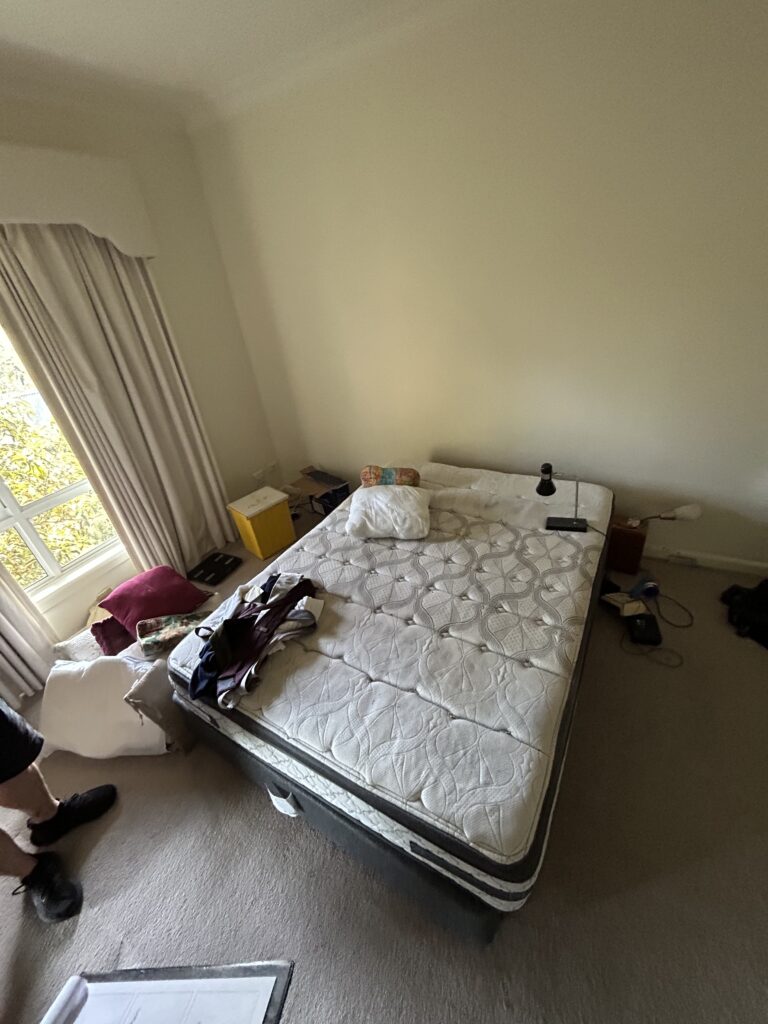 Unmade bed with clutter on mattress in a small sunlit bedroom, including a yellow bin and a nightstand by the window.