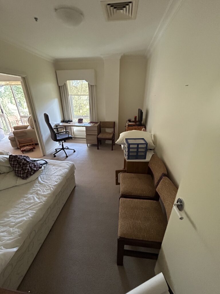 Hotel room with a bed on the left, a desk and swivel chair by a window, and three brown chairs along the right wall.