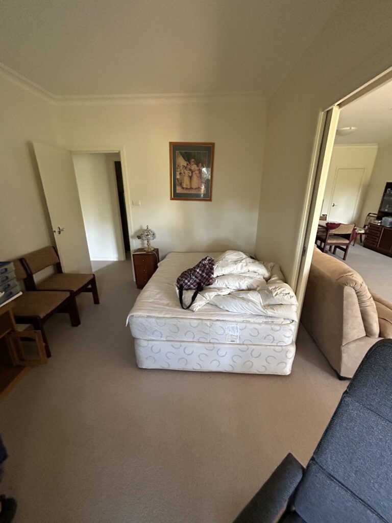 A messy bedroom with a placed twin mattress on the floor, covered by a quilt and a plaid bag on top near a small nightstand lamp and framed painting on the wall.