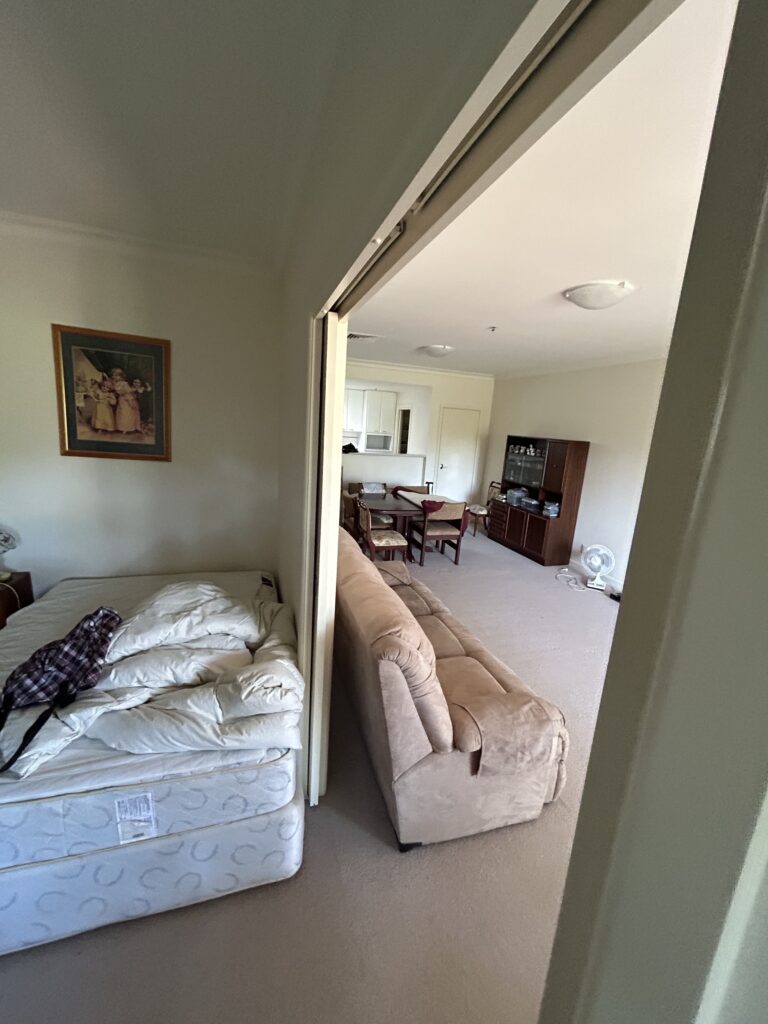 Bedroom with an unmade mattress on the floor next to a beige couch, opening into a living area with a dining table and cabinet in the background.