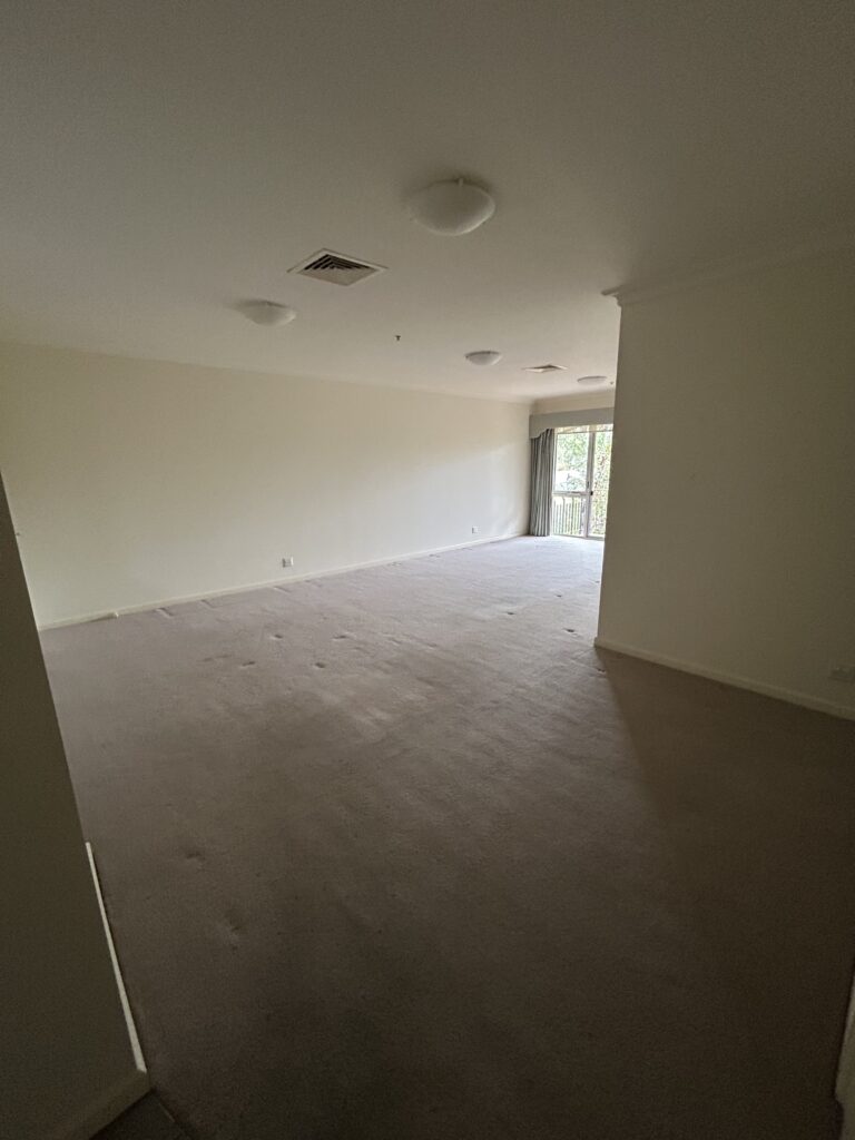 Empty living room with beige walls, light carpet, and ceiling lights, leading to a sliding glass door and balcony at the far end.