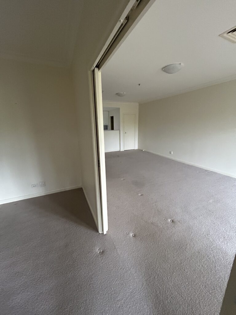 Empty living area with beige walls and a plush carpet, viewed from a doorway toward a small kitchen area in the back left.