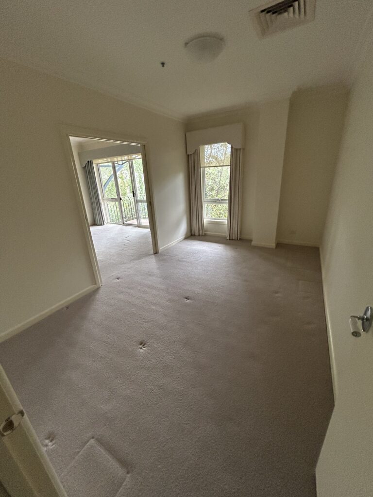 Empty beige-carpeted living room with a large window and doorway to a balcony area, light curtains, and an overhead ceiling light.