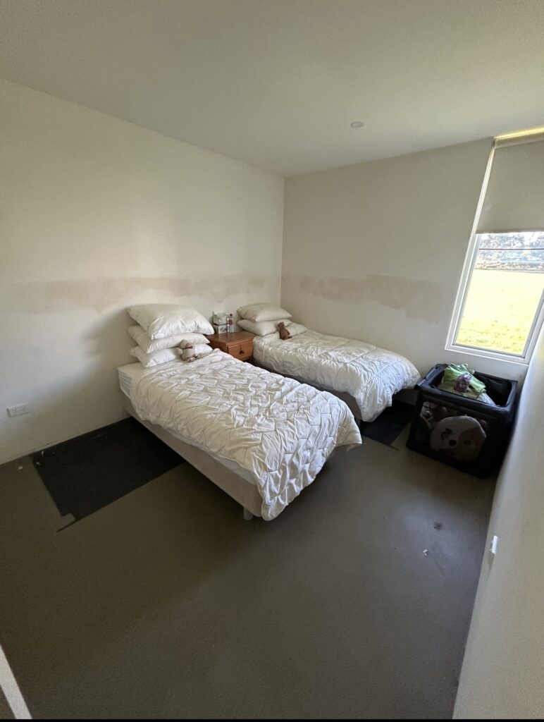 Small bedroom with two single beds, white quilts and pillows, and a shared bedside table between them; window to the right.