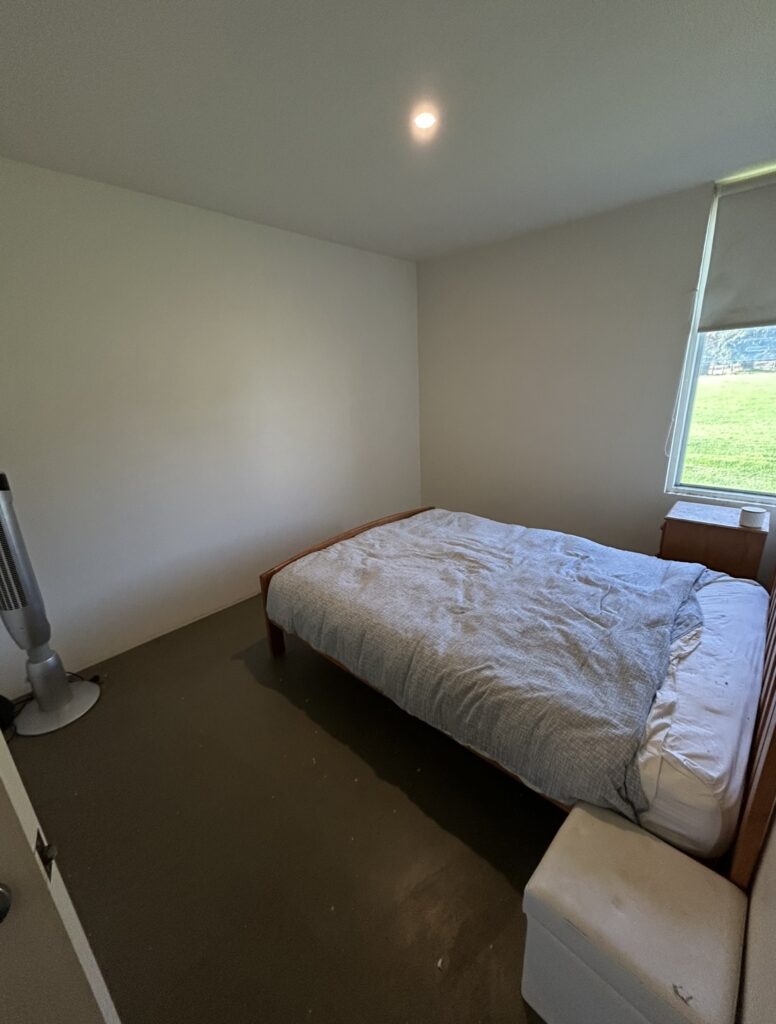 A small bedroom with a wooden double bed covered in a gray duvet, bare walls, and a window on the right letting in light. A tall fan stands in the corner.