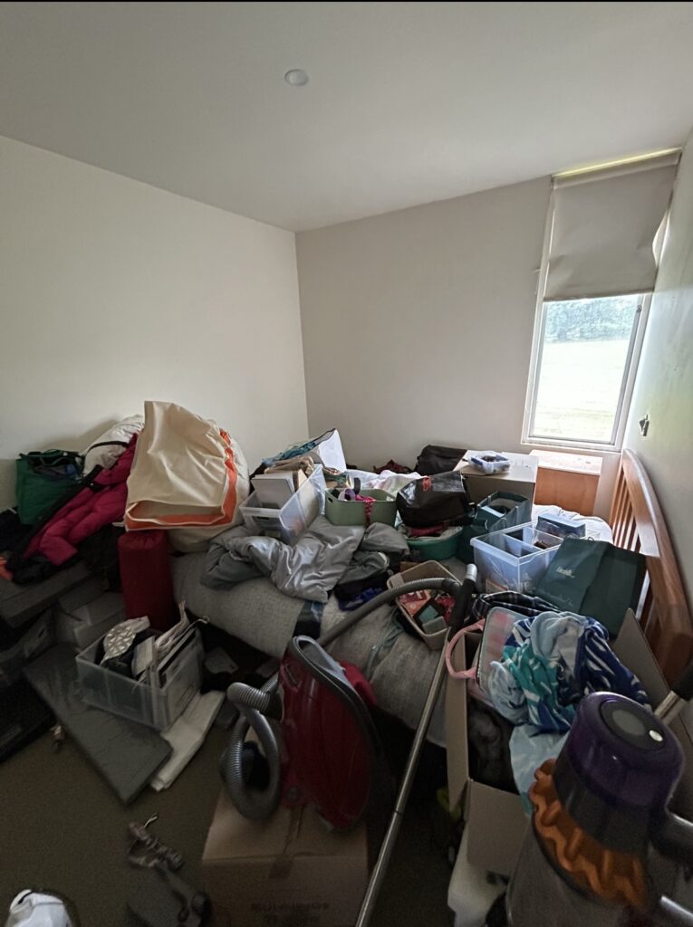 Messy bedroom with piles of clothes, bags, and storage bins piled on a bed and cluttered floor near a window.