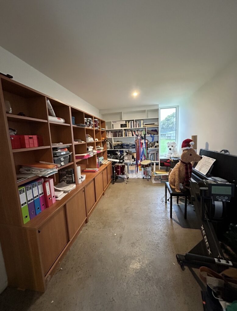 Cluttered home office/music room with tall wooden shelves of supplies, a drum set in the back, and a piano with a Santa-hat teddy on the bench