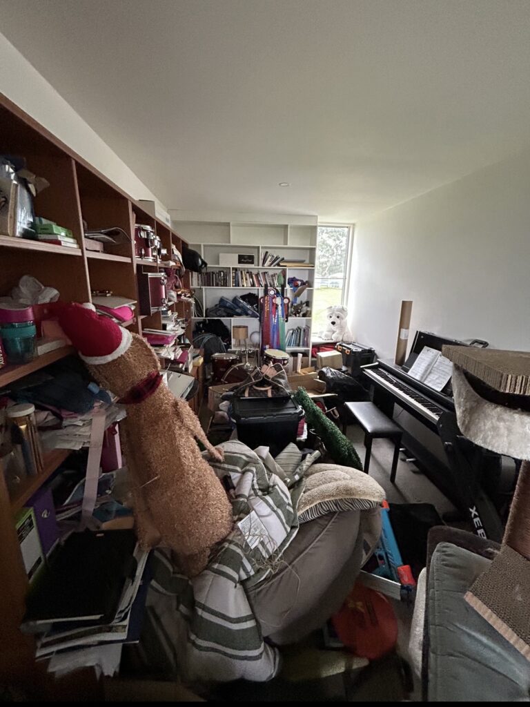 Very cluttered room with a piano on the right and crowded shelves filled with books, toys, and clothes; a window at the far end.
