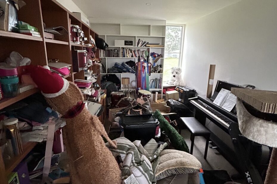 Very cluttered room with a piano on the right and crowded shelves filled with books, toys, and clothes; a window at the far end.
