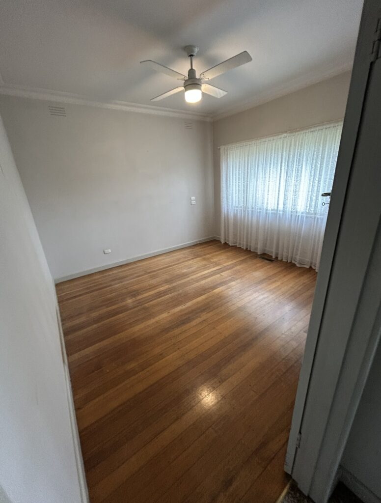 Empty room with hardwood floor, ceiling fan, and a large window with sheer lace curtains; door frame visible in the foreground.