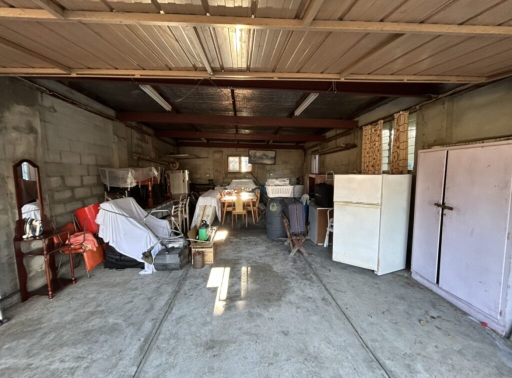 Cluttered garage with scattered furniture, appliances, and boxes along concrete floor and exposed ceiling beams