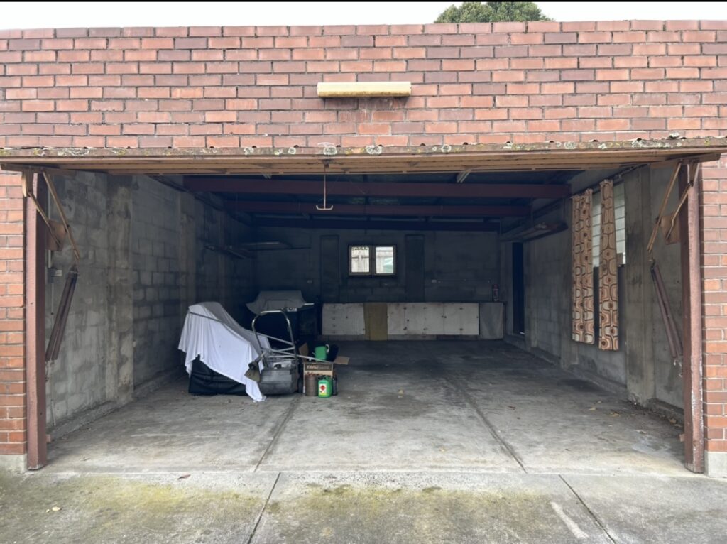 Open brick garage with a few stored items under a white tarp and scattered tools, a small window at the back, and a concrete floor.