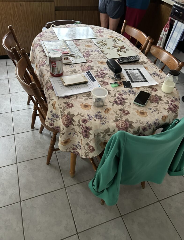 Round kitchen table with a floral tablecloth, cluttered with papers, a coffee can, a remote, phone, keys, and a green jacket on a chair