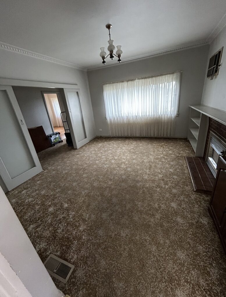 A sparsely furnished living room with beige carpet, a large window with sheer curtains, built-in white shelves, and a vintage chandelier overhead.