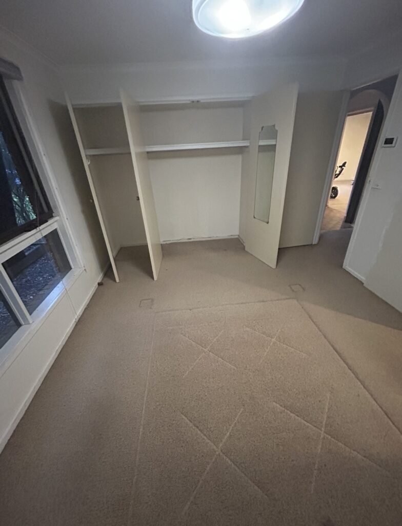 Empty bedroom with built-in wardrobes, doors open, mirror on the right closet, window to the left, beige carpet.