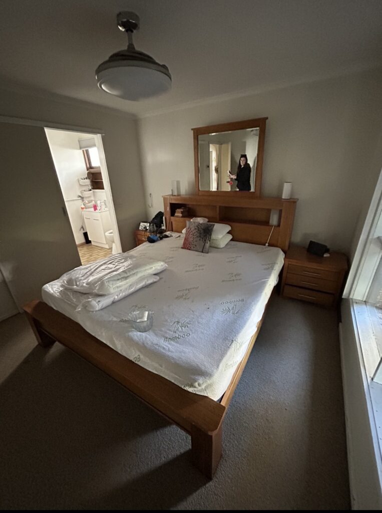 Wooden bed with white patterned sheets in a small bedroom; mirrors on the headboard, two nightstands, and an open bathroom door to the left.
