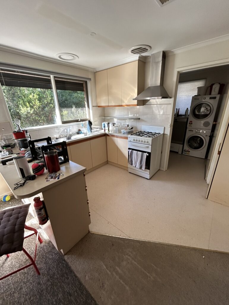 Bright L-shaped kitchen with beige cabinets, a stainless steel range hood, and a gas stove; laundry appliances visible in the adjoining room to the right.