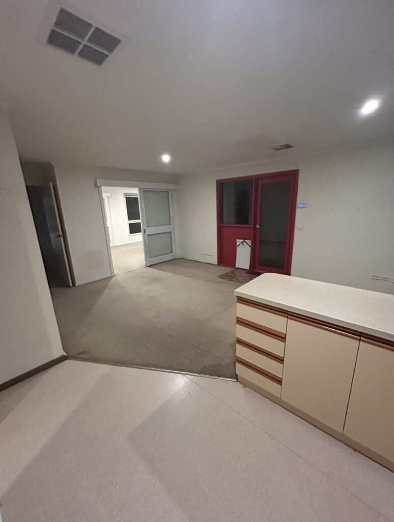 Empty living area in an apartment: white walls, beige carpet, red-framed door with glass panels, and a white kitchen counter on the right; doorway to another room ahead.