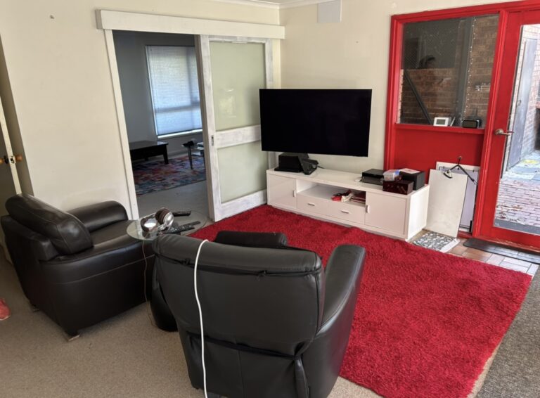 Living room with two black leather recliners, a white TV stand, and a large red area rug; red exterior door to the right and a doorway to another room on the left.
