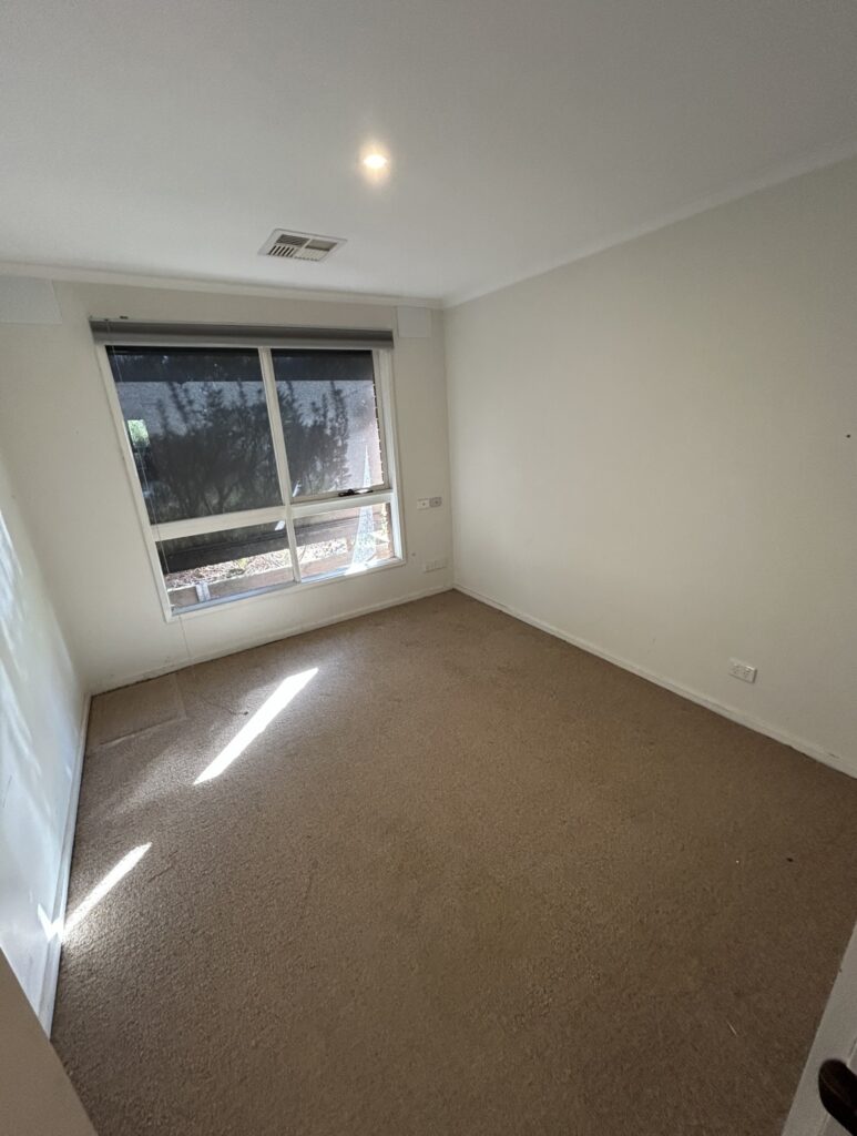 Small empty room with beige carpet, white walls, and a large window letting in sunlight.