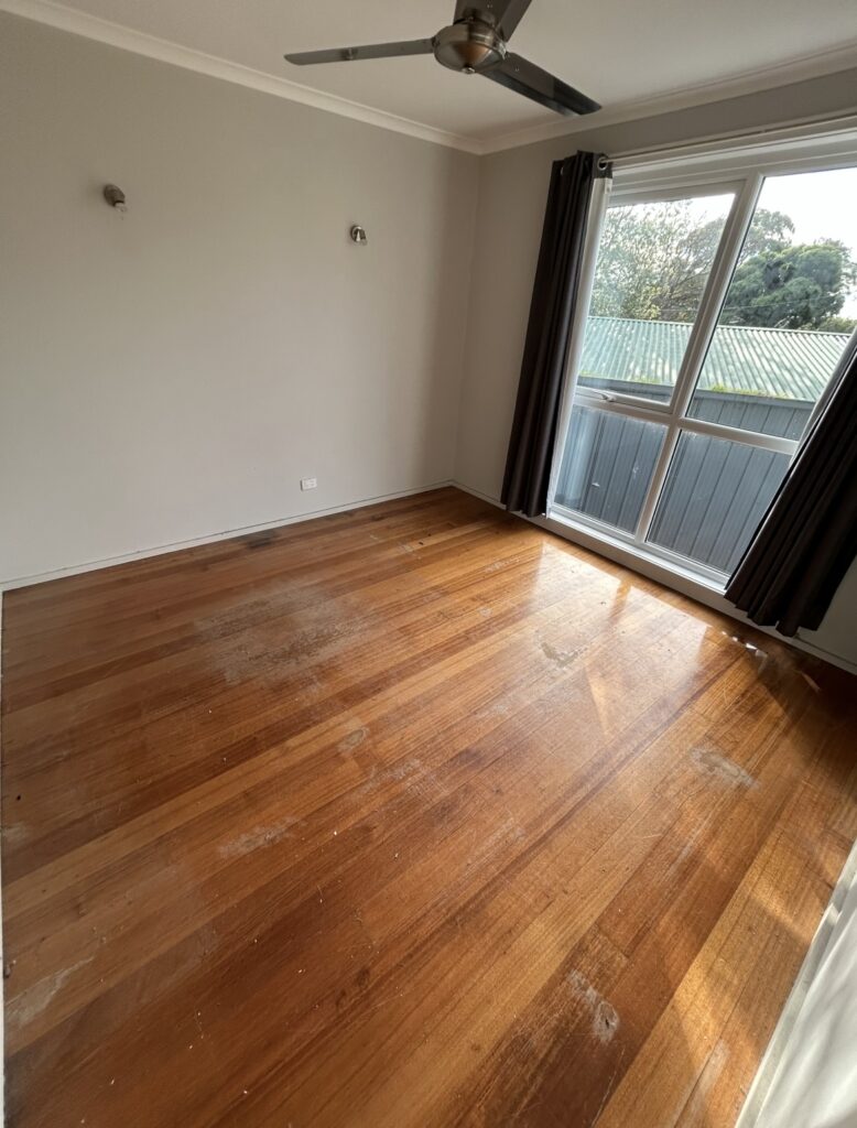 Empty room with light beige walls, hardwood floors, a ceiling fan, and a large window with dark curtains looking out to a fence and greenery.