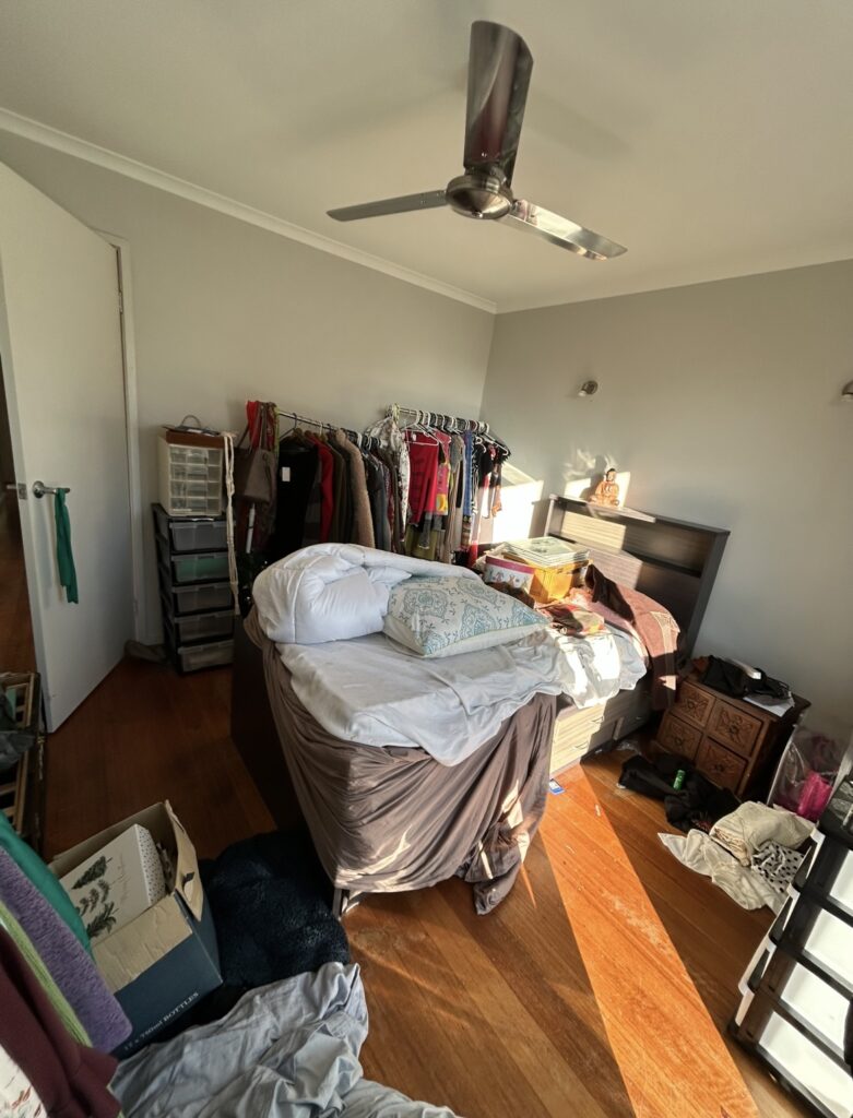 Messy bedroom with a cluttered bed, clothing rack, drawers, and various belongings on the floor and dresser.