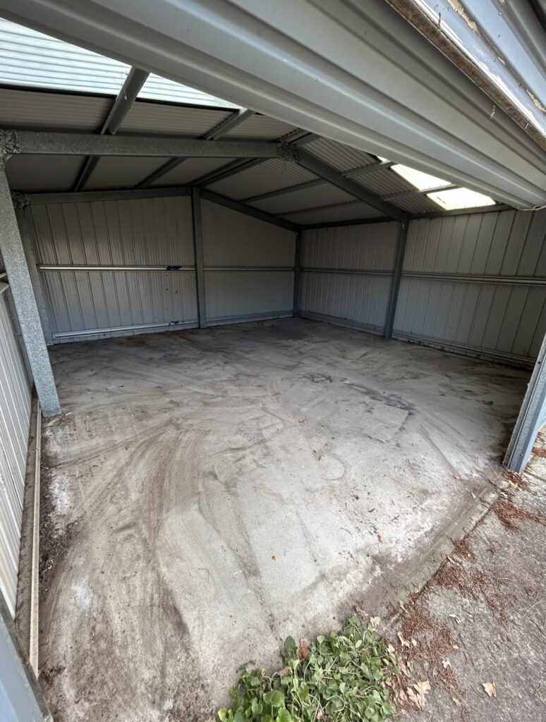 Empty metal storage shed interior with corrugated steel walls, exposed beams, and a bare concrete floor seen from the doorway, with a small plant at the threshold.