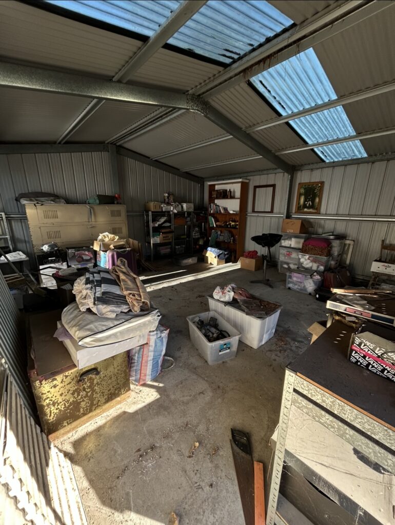 Cluttered metal garage storage room with shelves, boxes, and miscellaneous items scattered on the floor.