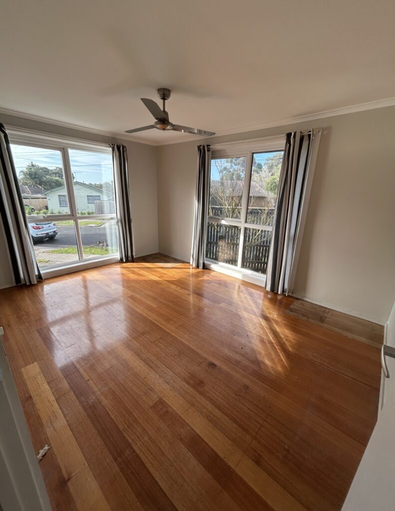 Bright empty living room with two large windows, striped curtains, and a silver ceiling fan, sunlight on the hardwood floor.