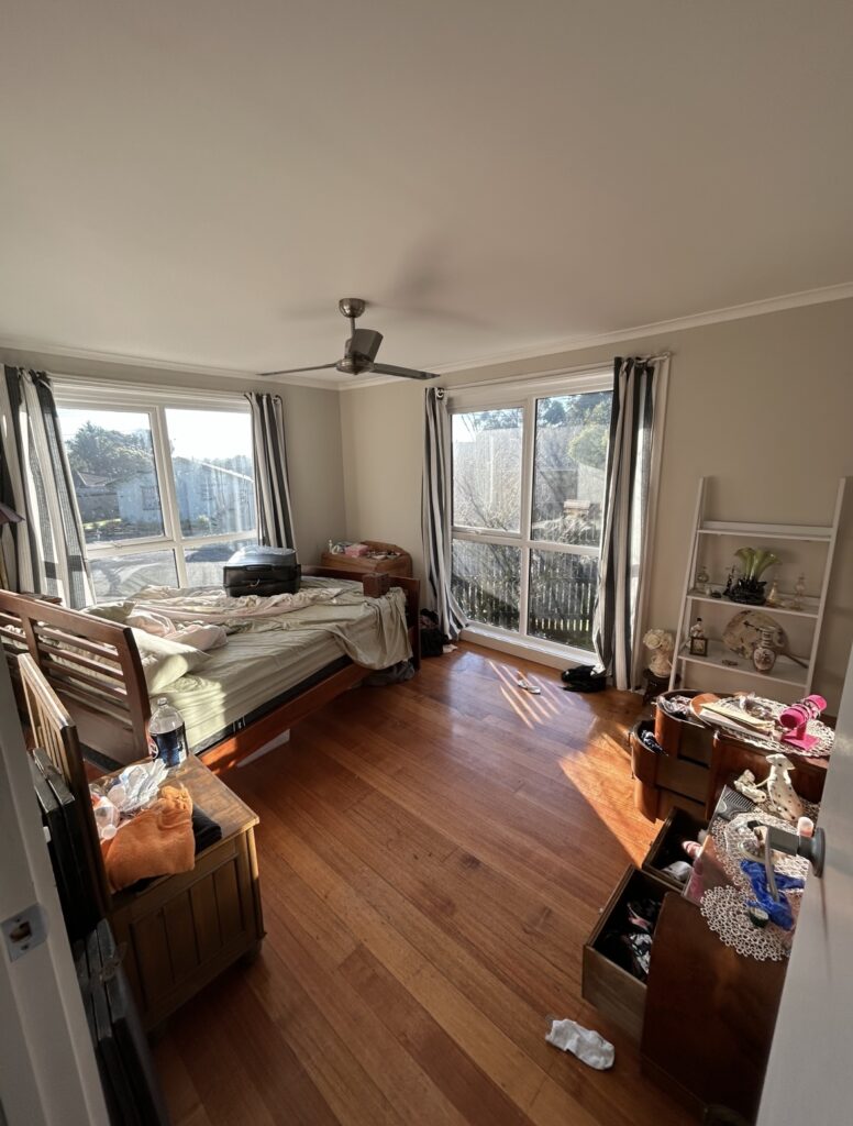 A small bedroom with large windows, an unmade bed, and cluttered surfaces bathed in afternoon light.