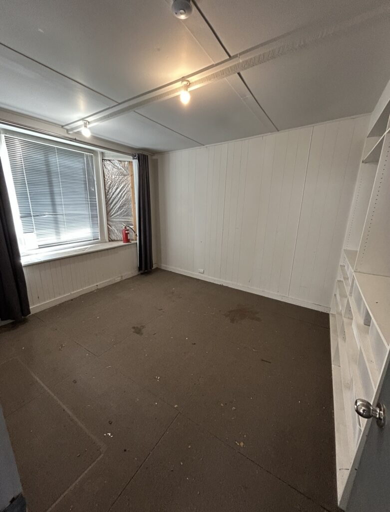 Empty, unfinished room with white paneled walls, brown carpet, and a window with blinds and dark curtains; a red fire extinguisher sits on the window ledge and built-in shelves are on the right side.