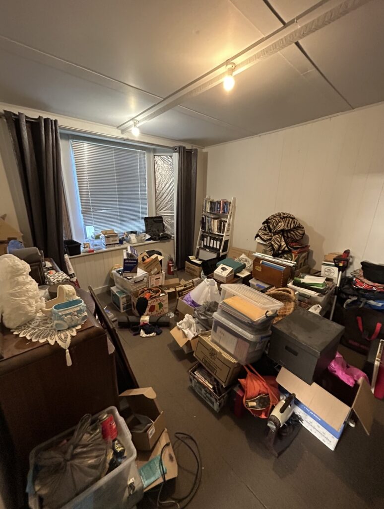 A cluttered storage room with piles of boxes, bins, and miscellaneous items scattered on the floor and furniture.