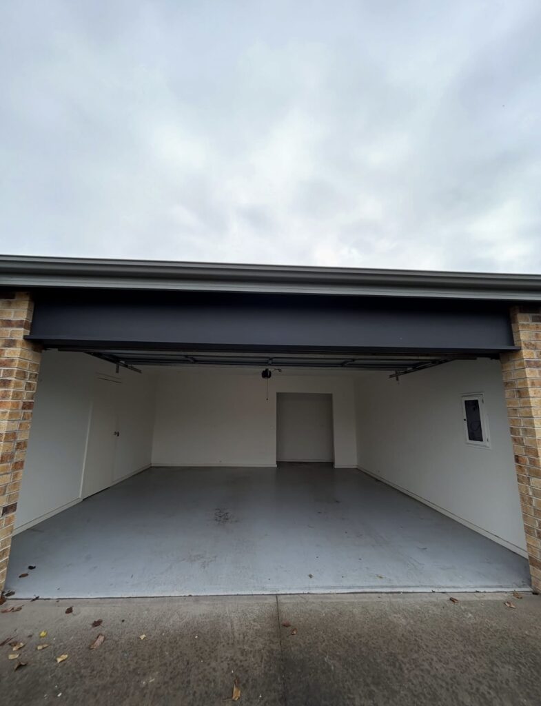 Empty two-car garage with white walls, gray concrete floor, brick sides, and a window on the right wall.