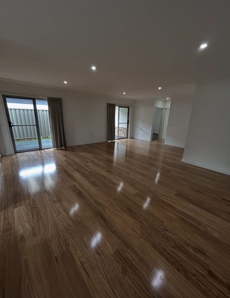 Bright, empty living room with polished hardwood floors, white walls, and recessed ceiling lights; sliding glass doors with curtains open to a small outdoor area.