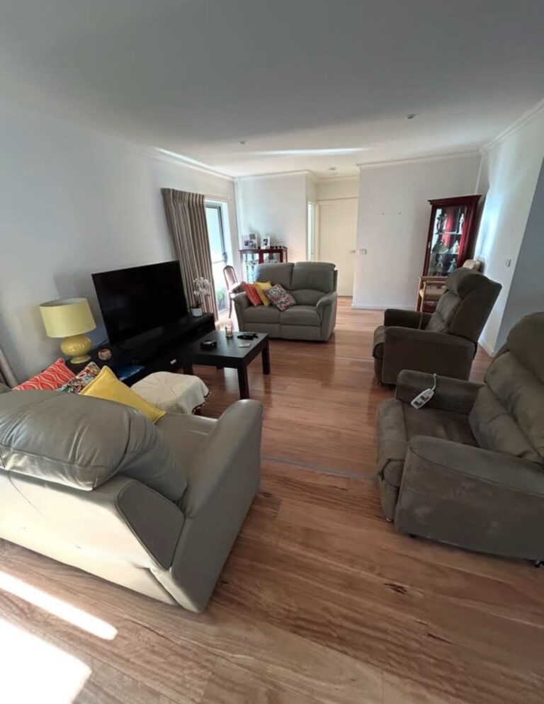 Living room with gray recliner sofas facing a large flat-screen TV, a black coffee table, and colorful pillows on a wooden floor.09A cozy seating area with a lamp and cabinet in the background.