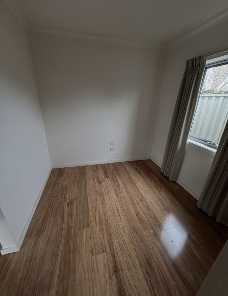 Empty small bedroom with light wood laminate floor, white walls, and a window with beige curtains.