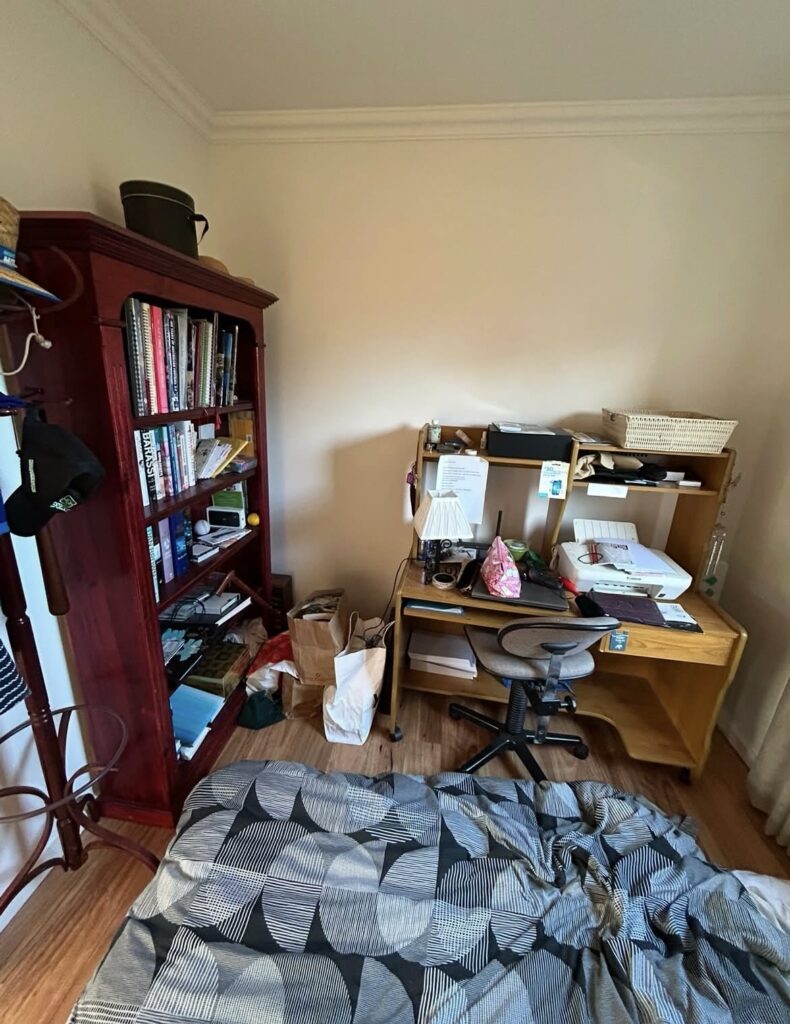 A small home office corner with a wooden desk, bookshelf, and cluttered surfaces, a chair, and a bed in the foreground; papers and bags on the floor.